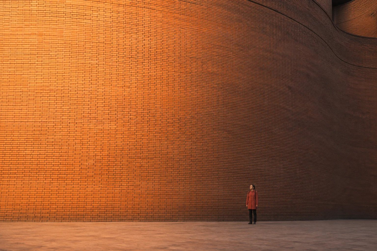 Small figure in a red coat against a monumental curved brick wall