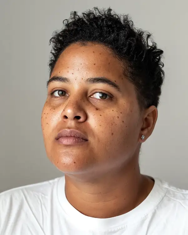 A woman with short curly black hair and a small earring, head slightly turned, in a white t-shirt against a grey background.