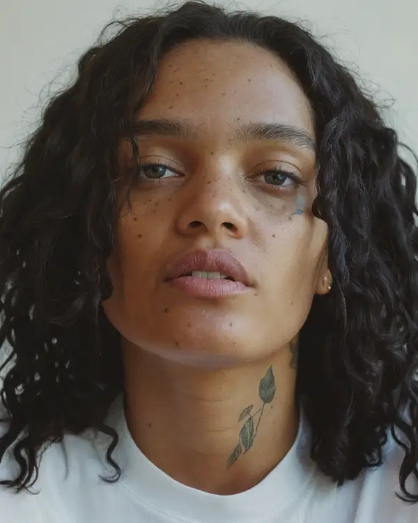A woman with long curly black hair, freckles and a small leaf tattoo on her neck, in a white t-shirt against a grey background.
