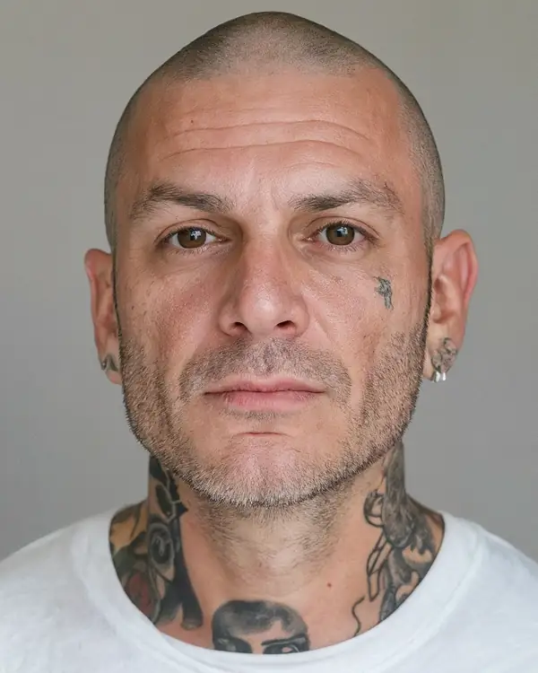 A man with a shaved head, small earrings and neck tattoos, in a white t-shirt against a grey background, looking at the camera.