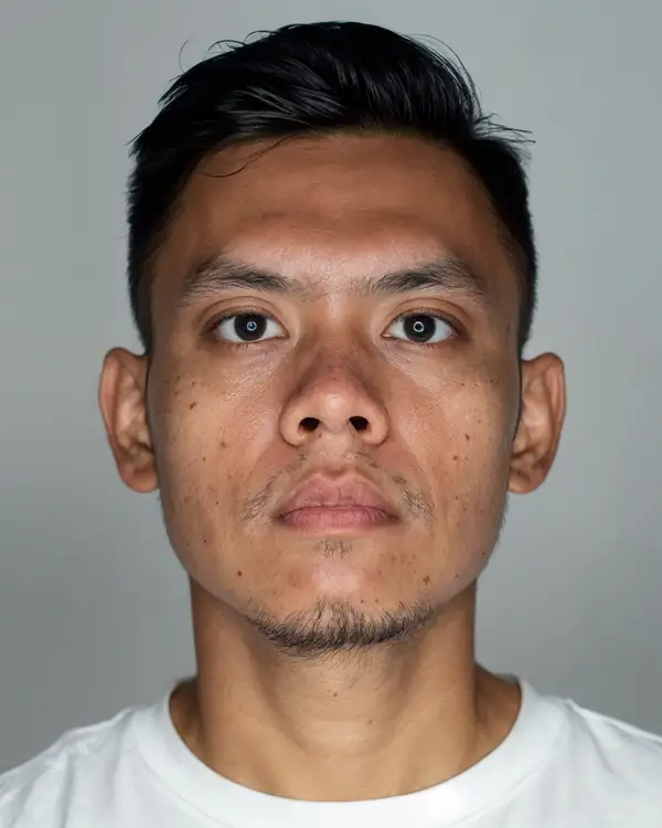 A young man with short black hair swept up, in a white t-shirt against a grey background, looking at the camera.