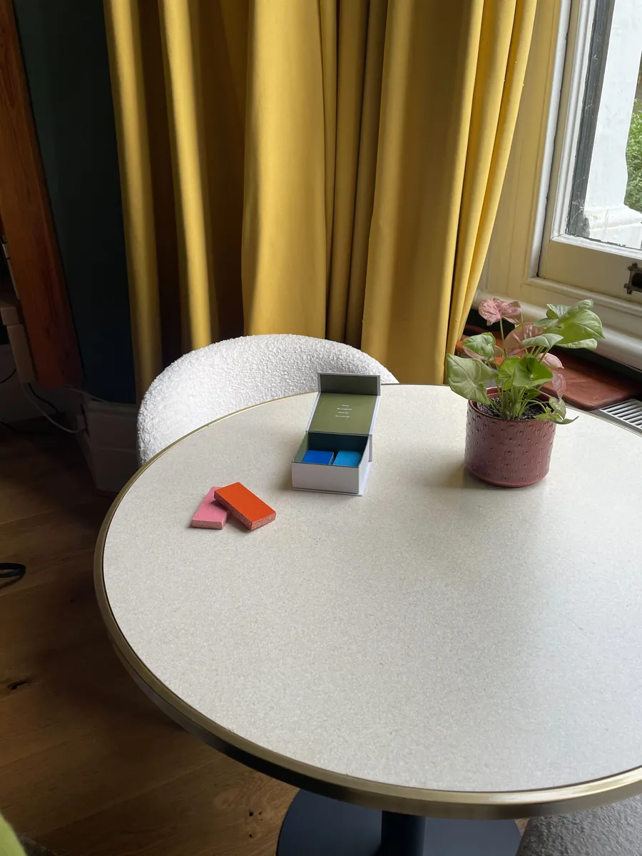 Physical swatch kit on a cafe table: open box of blue samples, loose pink and orange tiles, a small plant.