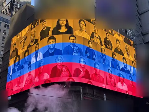 A curved corner screen in Times Square shows rows of portraits in yellow, blue and red, with street steam below.